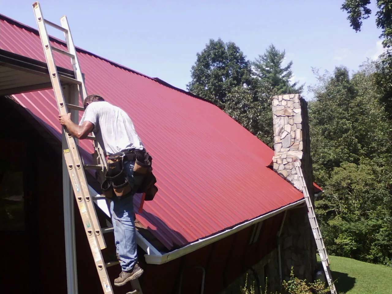 Roofer on a ladder beside a red metal roof and stone chimney — Soffit & Fascia Repair by Roofing Brothers in Rosemary Beach